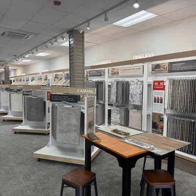 Interior of a carpet and flooring showroom in Duncan, OK, with multiple display racks of stone and tile samples, signage reading “CASUAL” and “TIMLESS,” and a small table with brochures in the foreground.